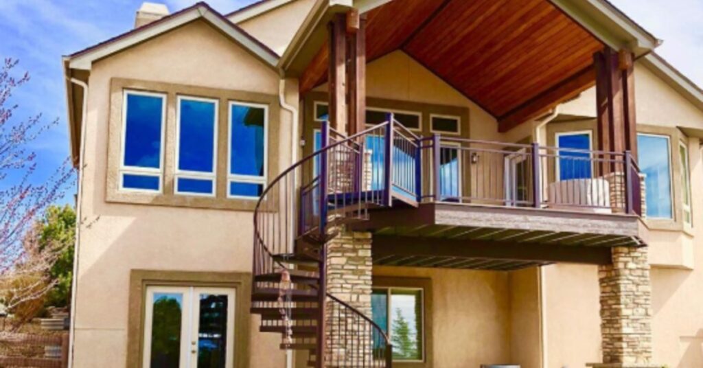 A two-story home with large windows, a covered balcony, stone columns, and a spiral staircase leading to the yard.