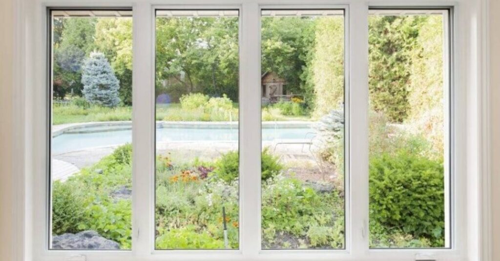 Large four-panel picture window overlooking a landscaped garden with shrubs, flowers, mature trees, and a pool.