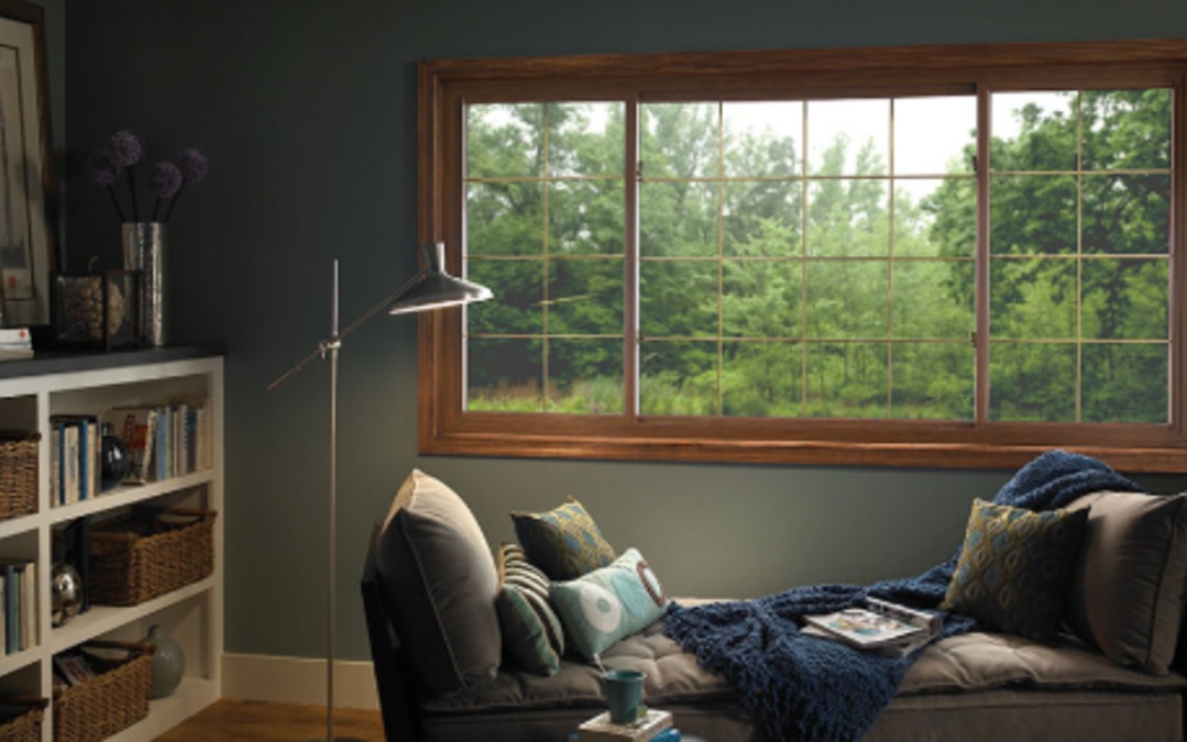 Cozy reading nook with a large wood-framed picture window overlooking lush green trees and filling the room with light.