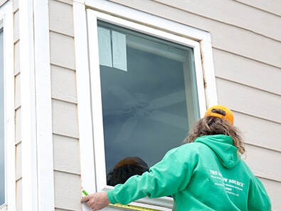 Worker in a green sweatshirt measures a white-trimmed window on a beige-sided home during an exterior project.