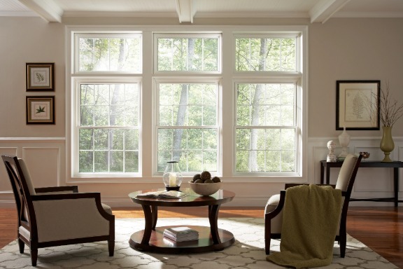 Bright living room with three large white windows, two armchairs, a round coffee table, and leafy trees visible outside.